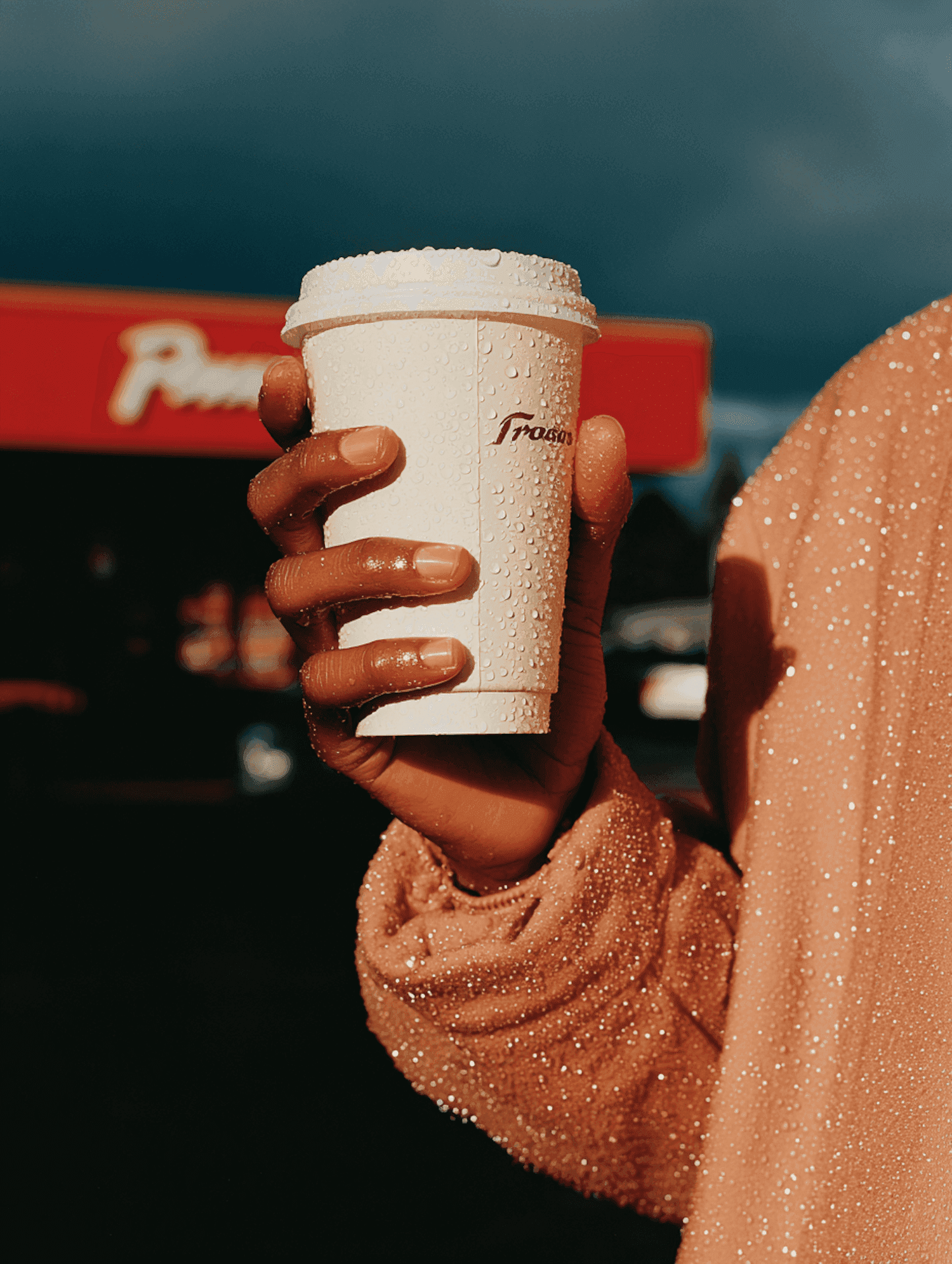 Hand holding a dewy coffee cup in front of a red storefront sign